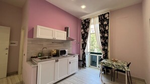 Cozinha compacta com armários brancos, micro-ondas e pia. Há uma área de refeições com mesa e cadeiras, além de uma janela com cortinas florais.