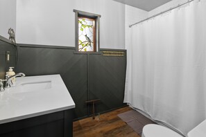 A bathroom with a white sink, a window with a bird decoration, a shower curtain, and a wooden floor.