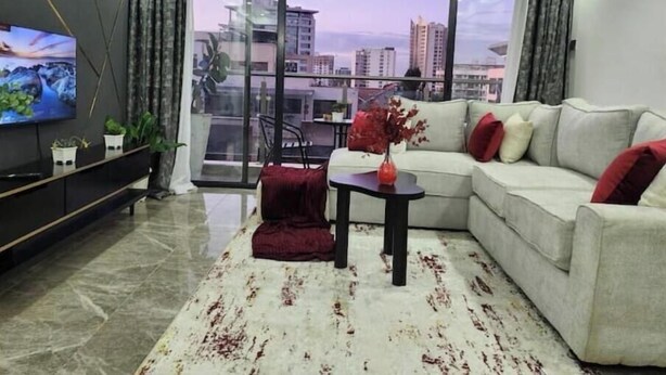 Una sala de estar moderna con un televisor grande, un sofá, una mesa de centro y vistas de la ciudad a través de amplias ventanas.