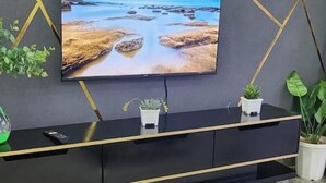 Una sala de estar moderna con un televisor de pantalla plana grande montado en una pared oscura, un mueble de entretenimiento negro con detalles dorados y plantas en macetas.