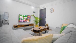 A modern living room with a grey sofa, a wooden coffee table, a flat-screen TV, a potted plant, and a clock on the wall. - Kilimani One bedroom (Nairobi)