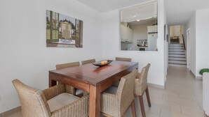A dining area with a wooden table and wicker chairs, a painting on the wall, and a kitchen visible through an open door. - Apartment 'Infinity View Menorca' with Sea View, Shared Pool and Private Terrace (Es Mercadal)