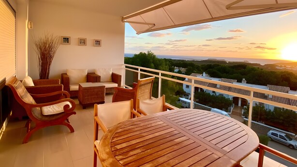 A balcony with wicker furniture, a wooden table, and a view of the sunset over a landscape. - Apartment 'Infinity View Menorca' with Sea View, Shared Pool and Private Terrace (Es Mercadal)