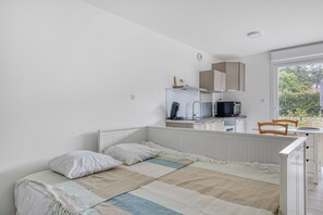 A bedroom with a bed, a bedside table, a chair, a window, and a kitchenette.