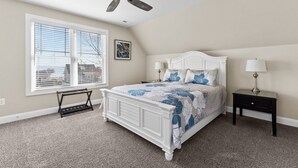 Une chambre à coucher avec un lit, un ventilateur de plafond, une table de chevet, une lampe, une fenêtre avec des stores et un tableau au mur.