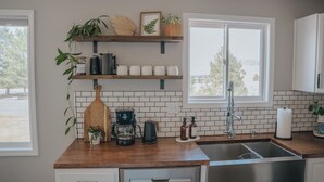 A kitchen with wooden countertops, a stainless steel sink, and a window with a view of trees. There are shelves with plants and kitchen items.