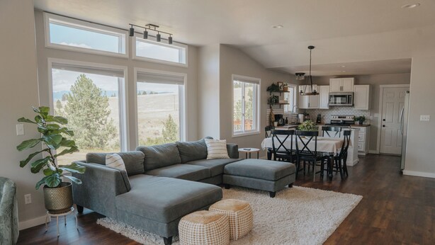 A spacious living room with a gray sectional sofa, a dining area with a black table and chairs, and a kitchen with white cabinets and a microwave.