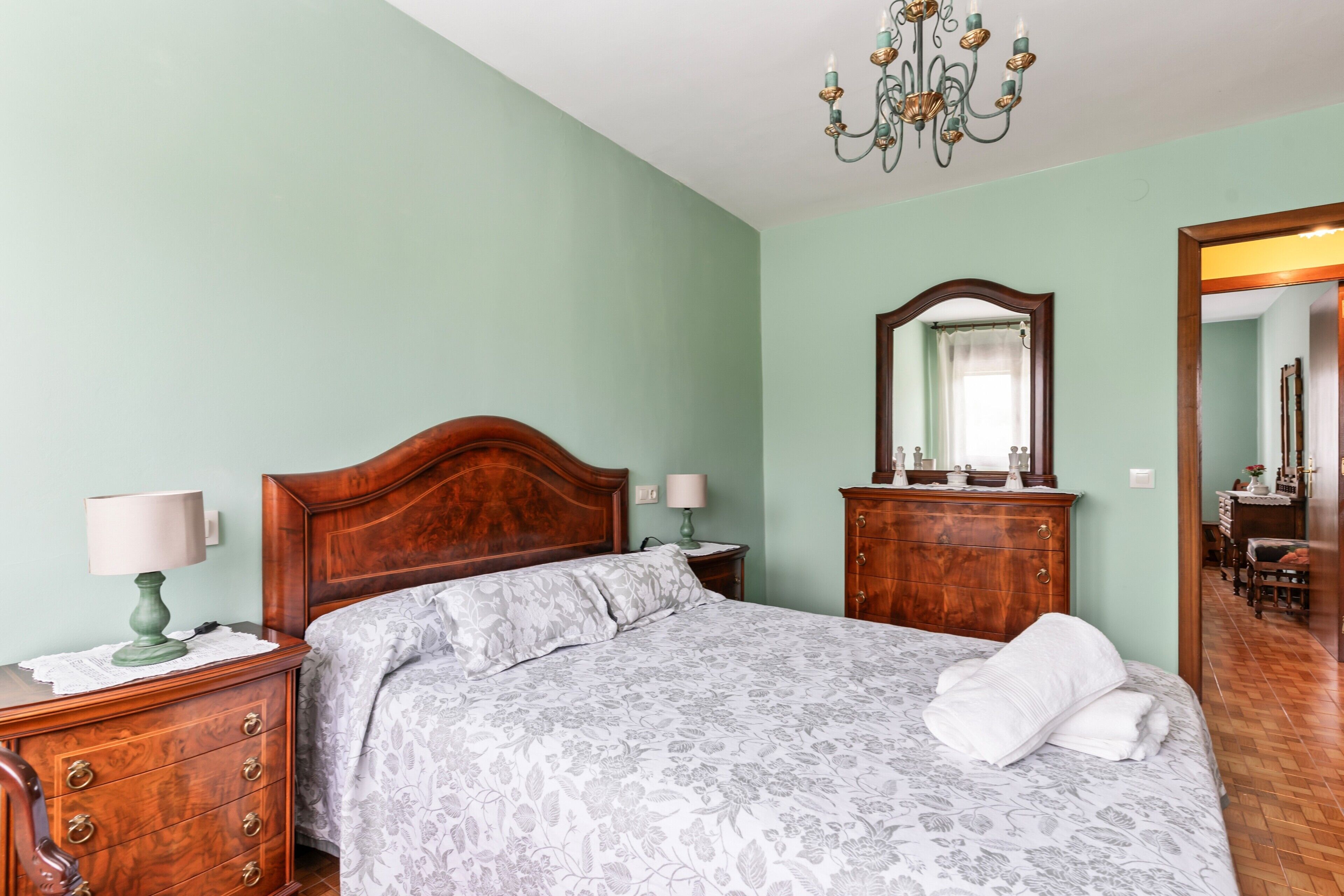 A bedroom with a wooden bed, two bedside tables, a dresser, a mirror, and a chandelier.