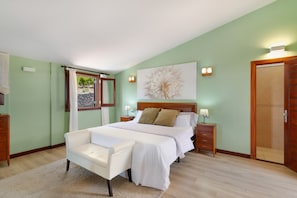 Quarto com cama, mesas de cabeceira, uma cadeira, janela com cortinas, uma obra de arte na parede e uma porta.