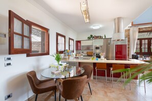 Cozinha moderna com ilha central, sala de jantar com mesa e cadeiras de vidro, e vista para o exterior.