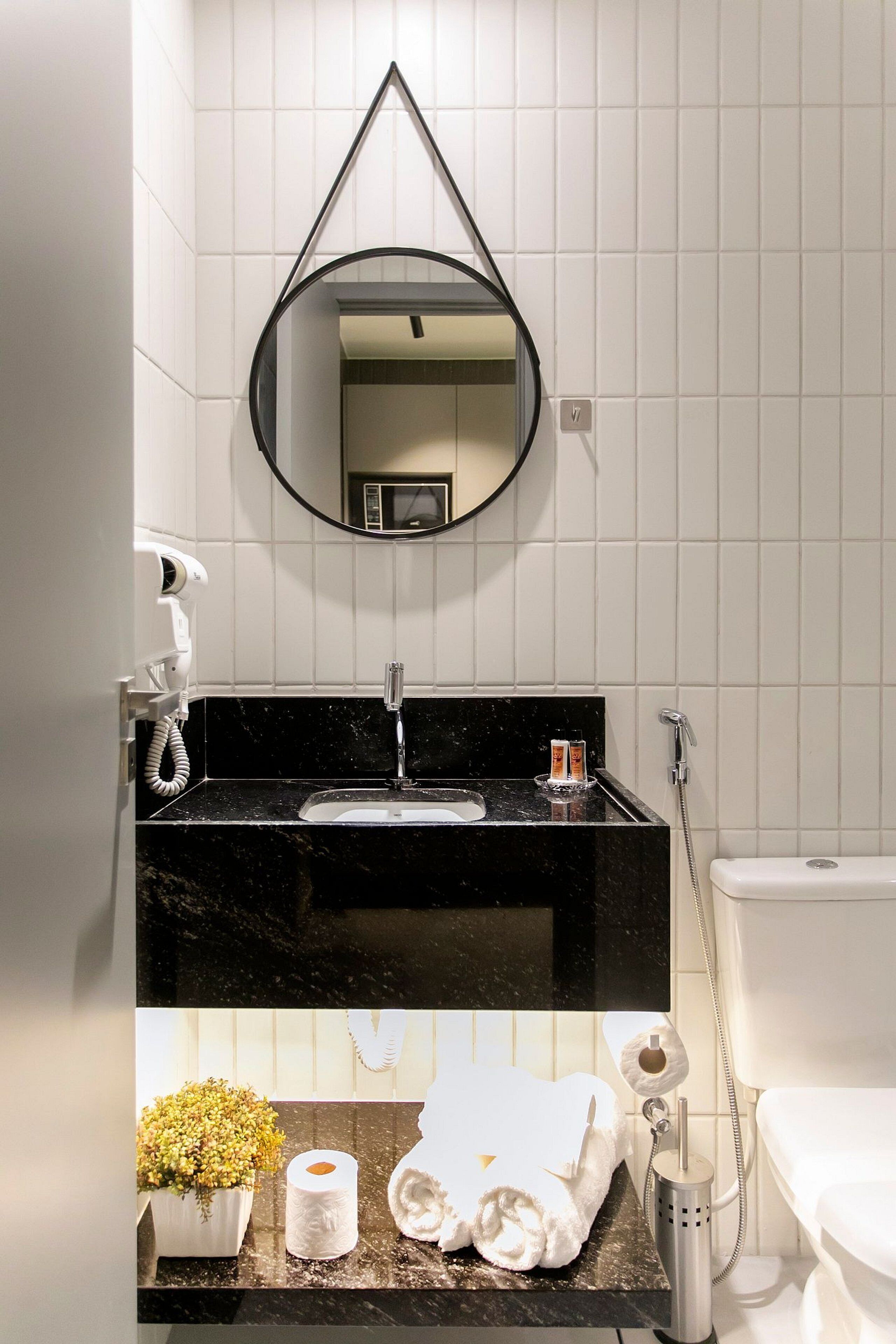 Casa de banho com lavatório preto, azulejos brancos, espelho, secador de cabelo, toalha e sanita.