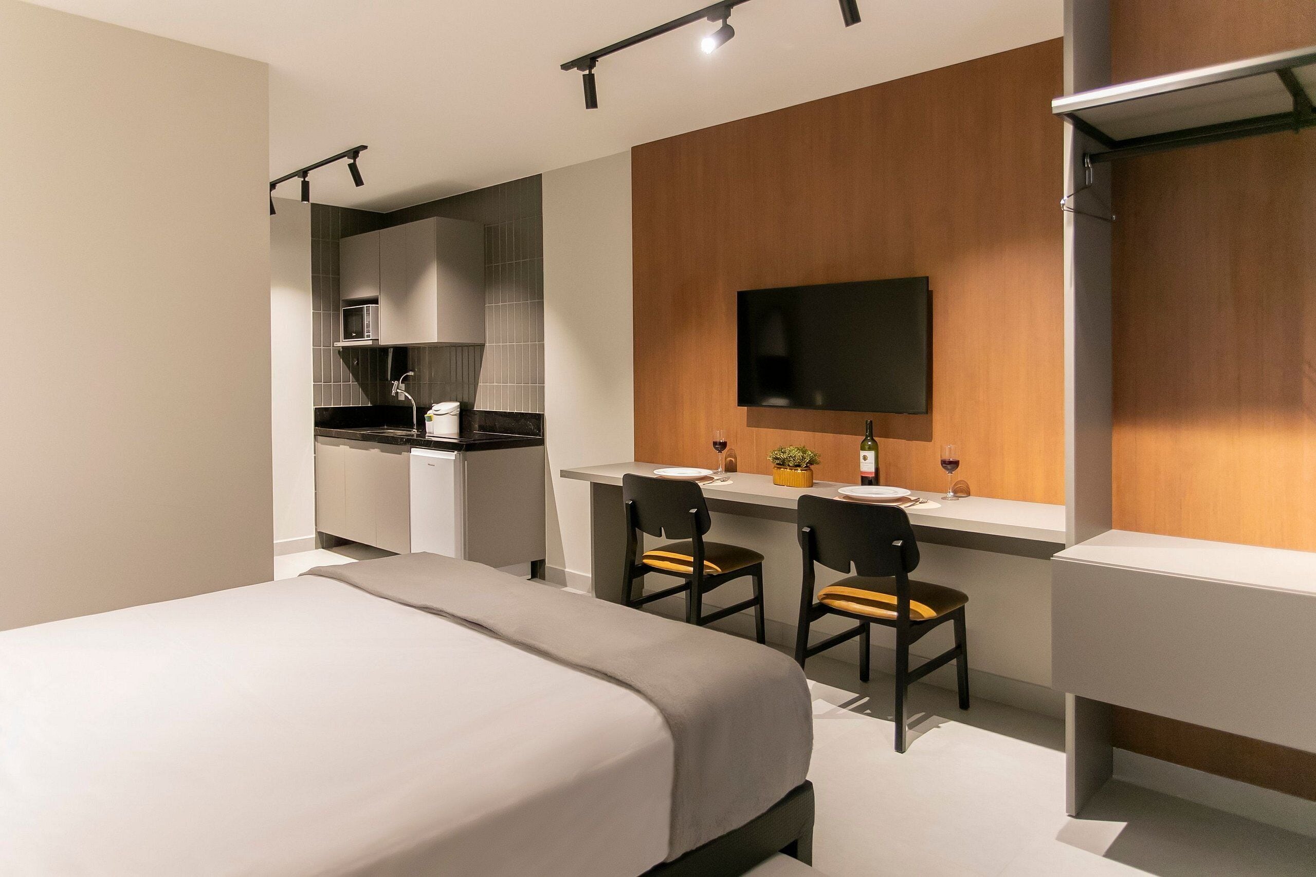 A modern hotel room with a bed, a desk with two chairs, a flat-screen TV mounted on a wooden wall, and a kitchenette in the background.