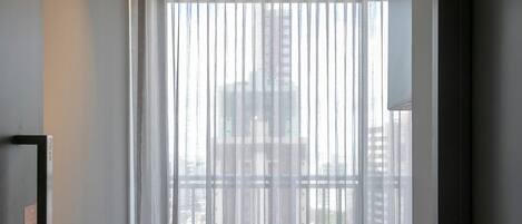 A modern hotel room with a large window, sheer curtains, and a ceiling-mounted light fixture.