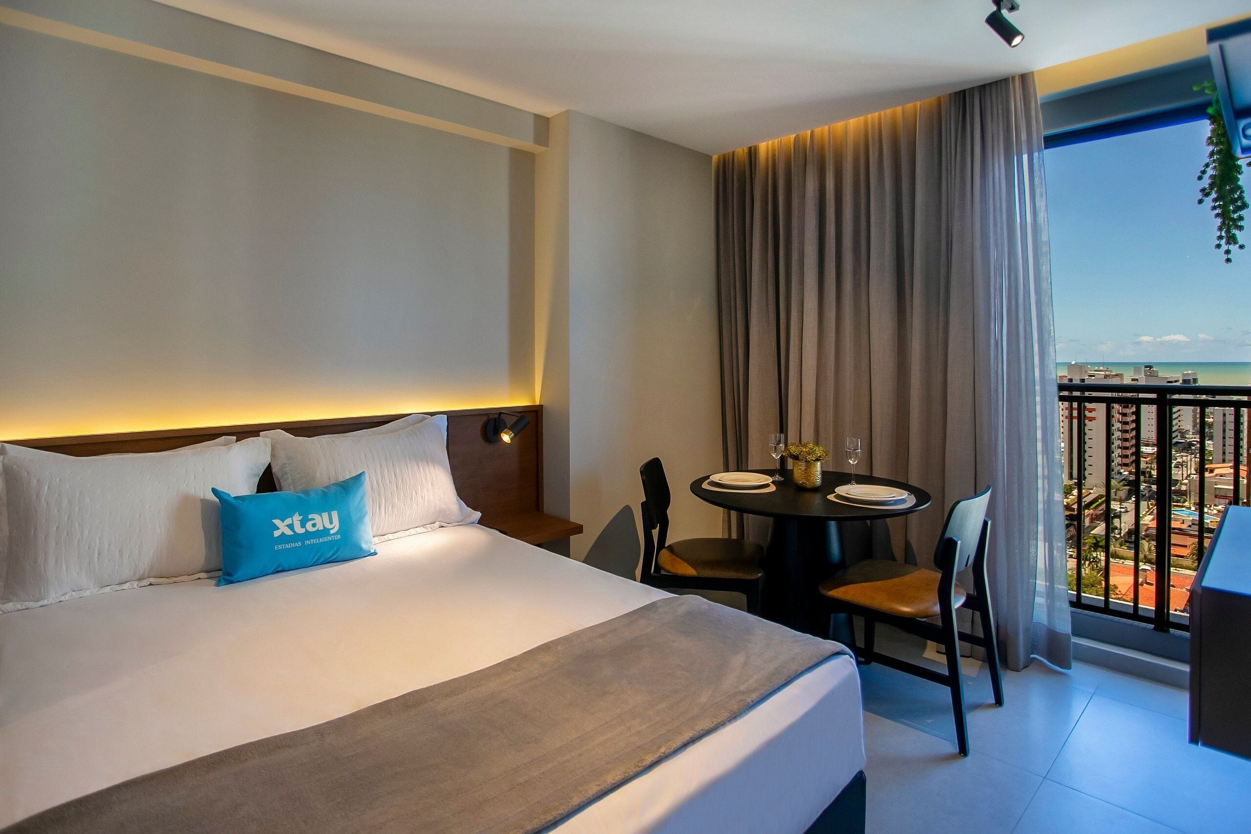 A hotel room with a bed, a table with chairs, a balcony with a view of the city, and a window with curtains.