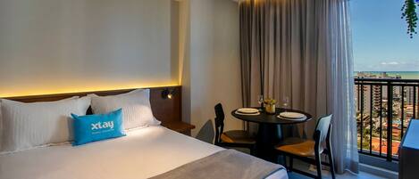 A hotel room with a bed, a table with chairs, a balcony with a view of the city, and a window with curtains.