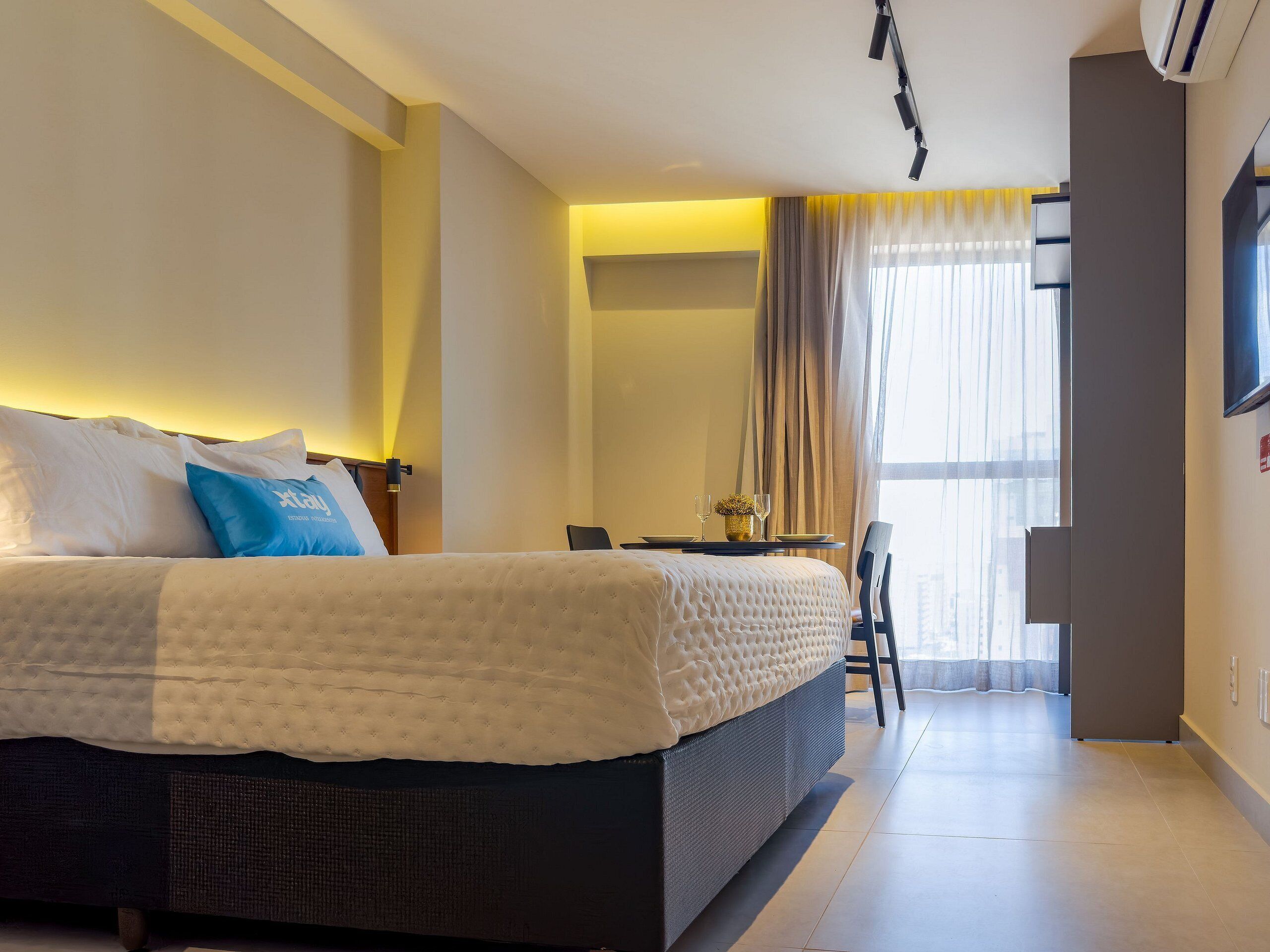 Een hotelkamer met een groot bed, een eettafel met stoelen en een aan de muur bevestigde televisie.