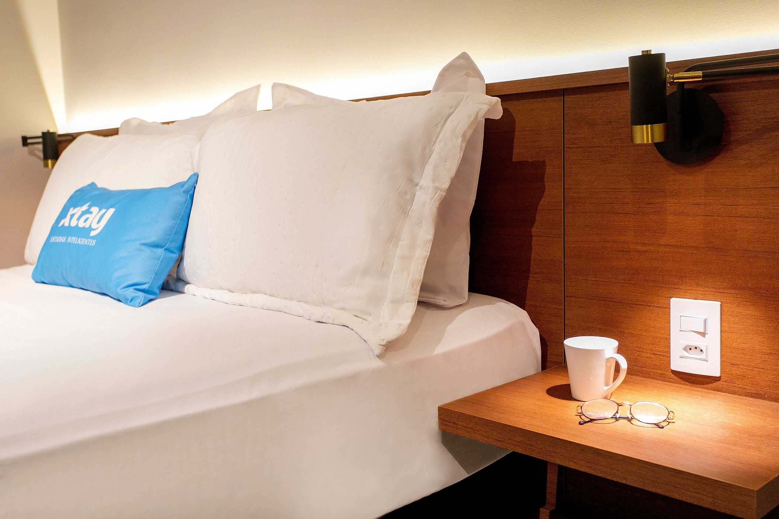 Una cama bien hecha con ropa blanca y una almohada azul, una mesita de noche con una taza blanca y vasos, y una lámpara fijada a la pared.