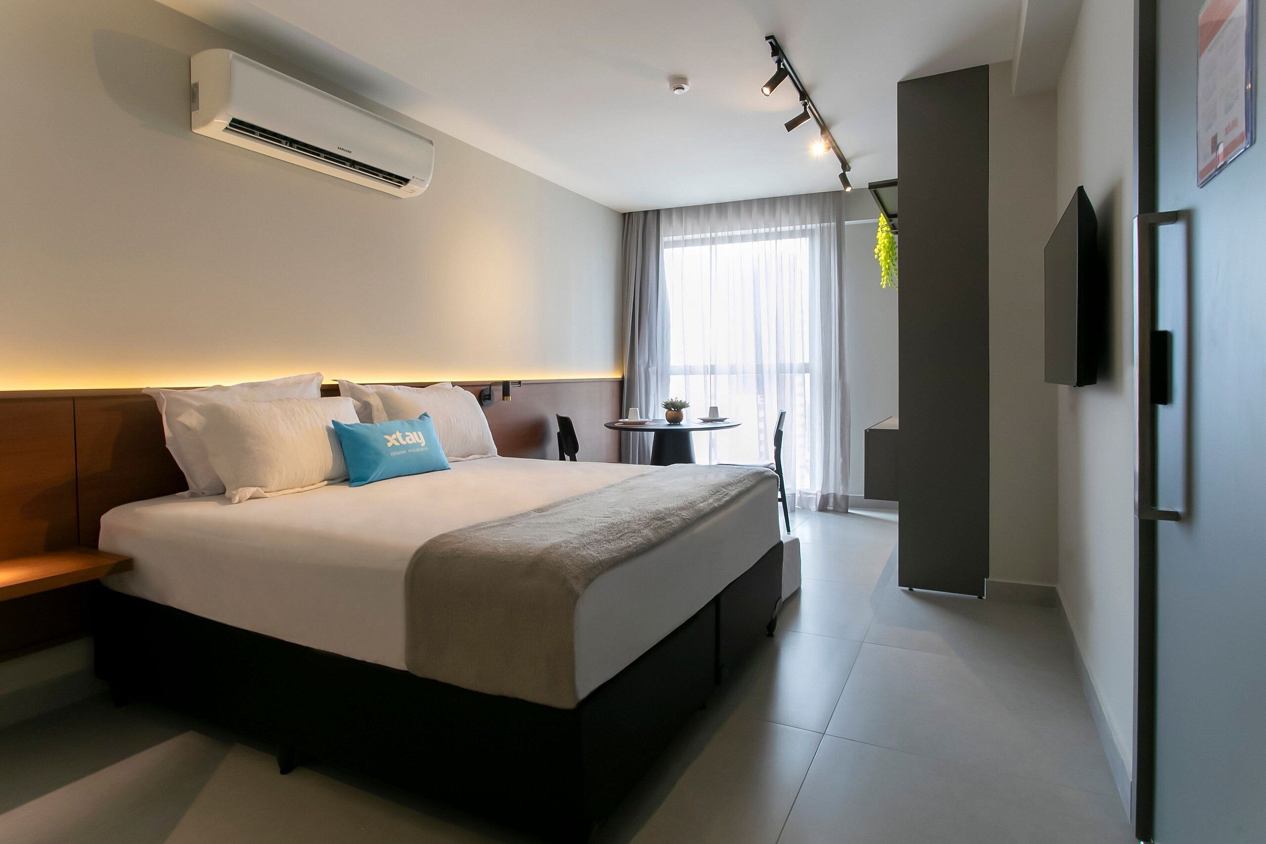 Une chambre d’hôtel moderne avec un grand lit, des tables de chevet, un appareil de climatisation, une petite table ronde avec une plante et un téléviseur fixé au mur.