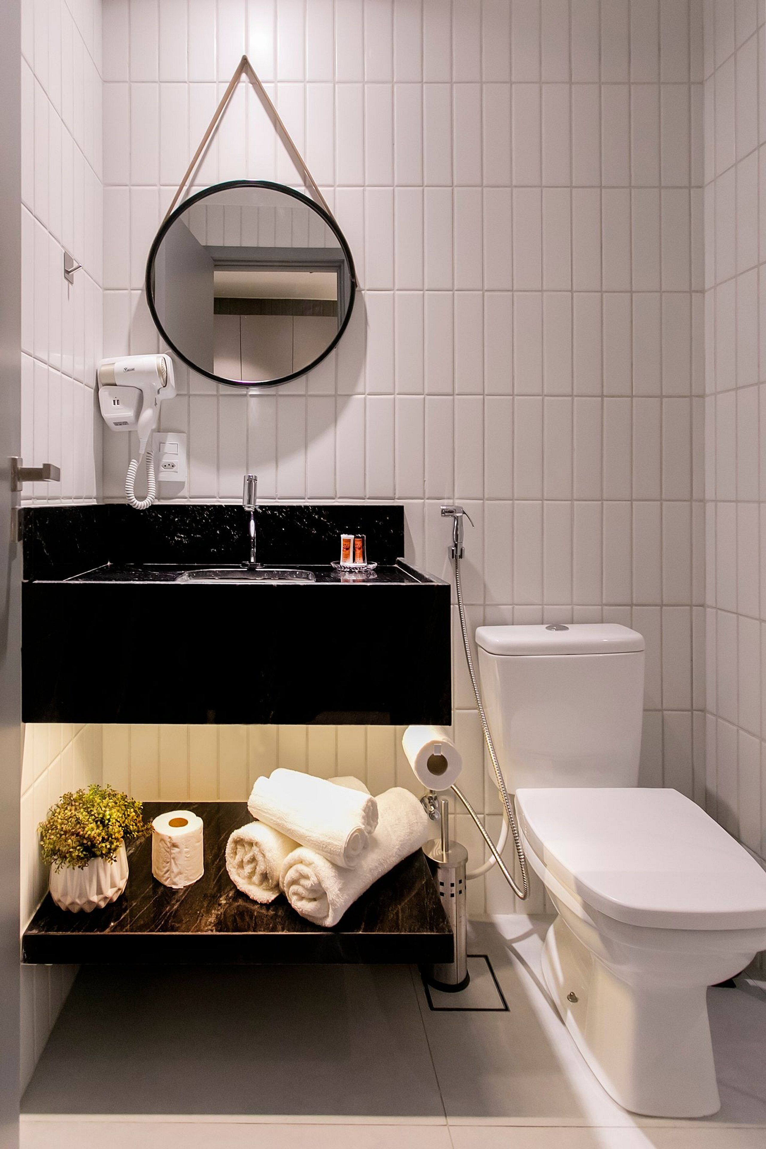 A modern bathroom with a black countertop, white tiles, a round mirror, a toilet, a hairdryer, and a small potted plant.