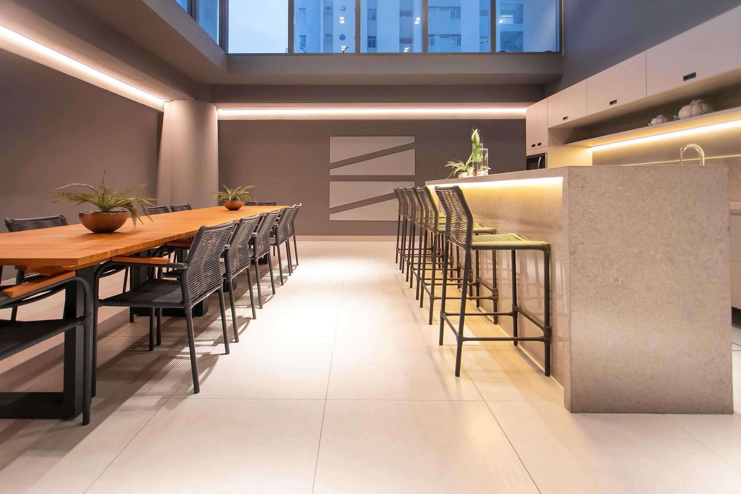 A modern dining area with a long wooden table and black chairs, a kitchen area with a marble island, and a view of the cityscape through large windows.