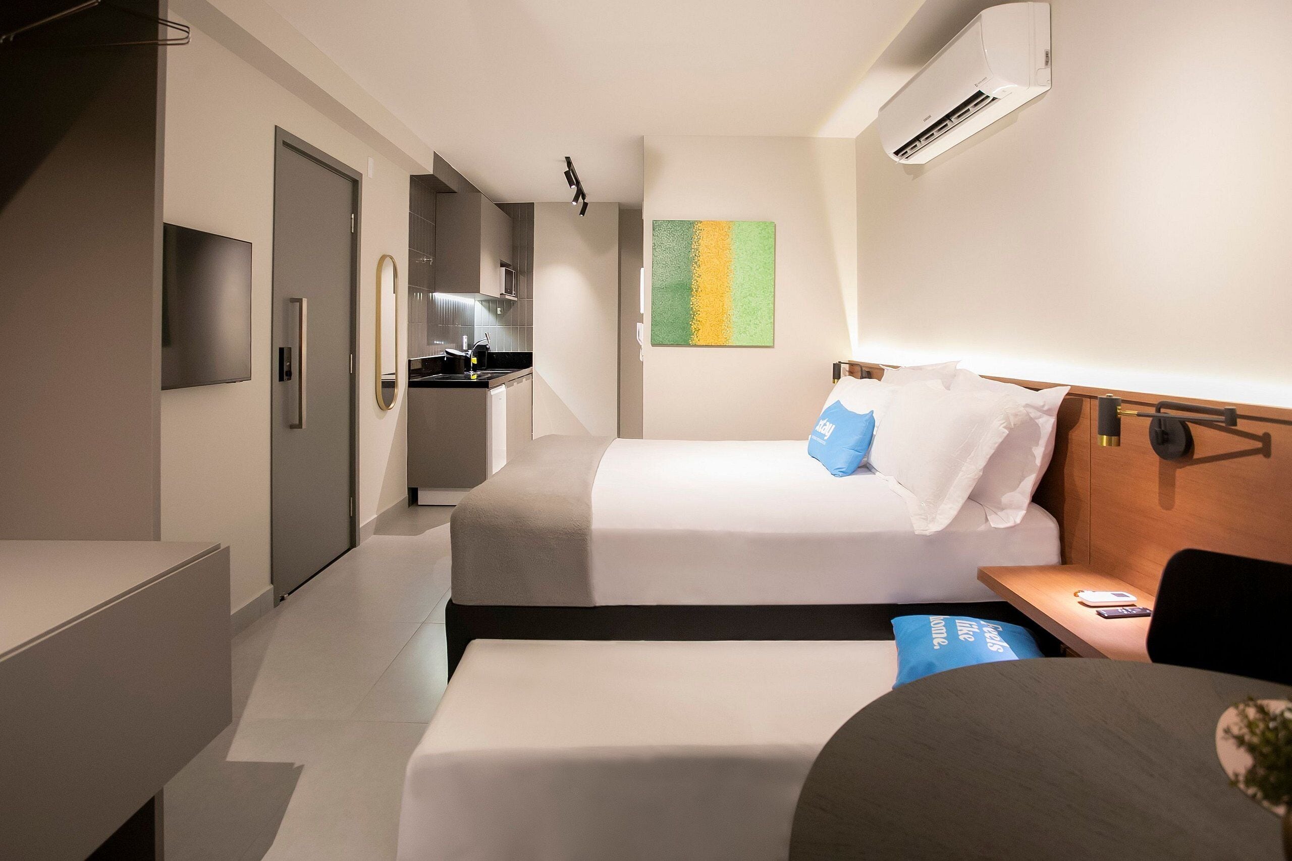A modern hotel room with a bed, bedside table, and a small desk. There is a kitchenette in the background.