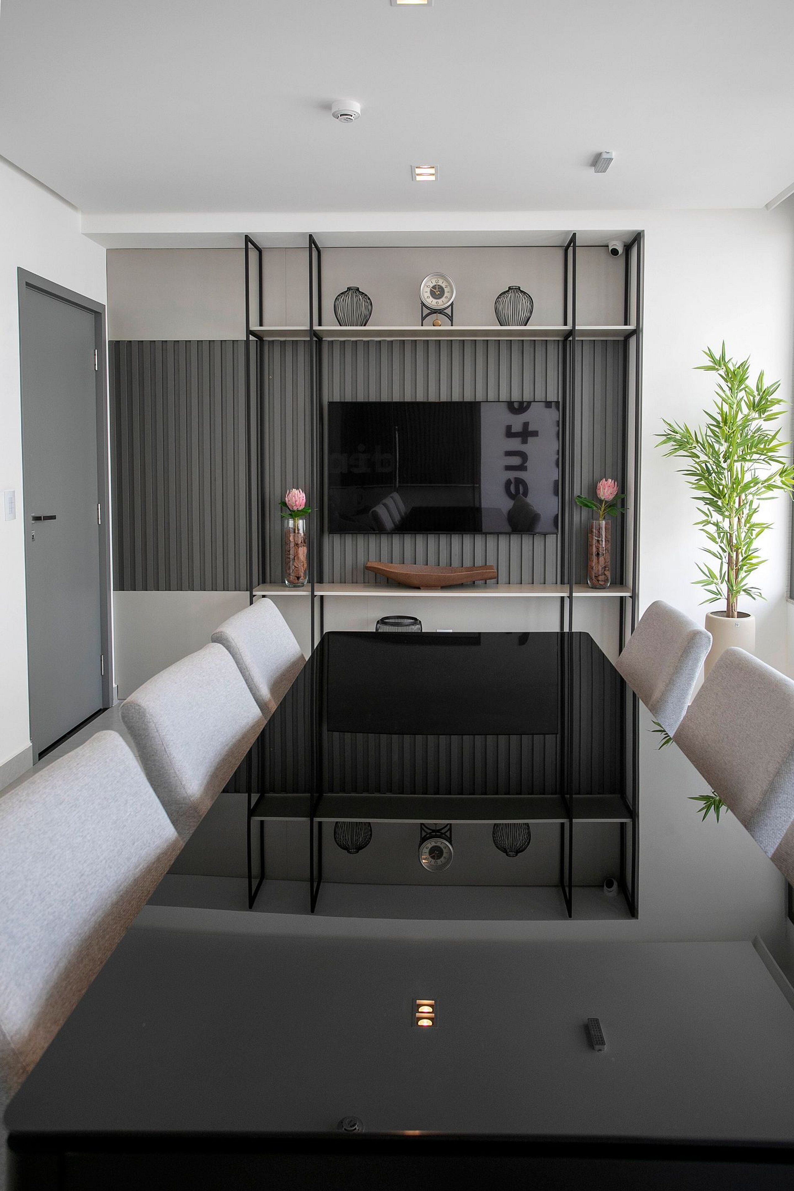 A modern living room with a large flat-screen TV mounted on a wall, a black coffee table, two grey sofas, and a potted plant.