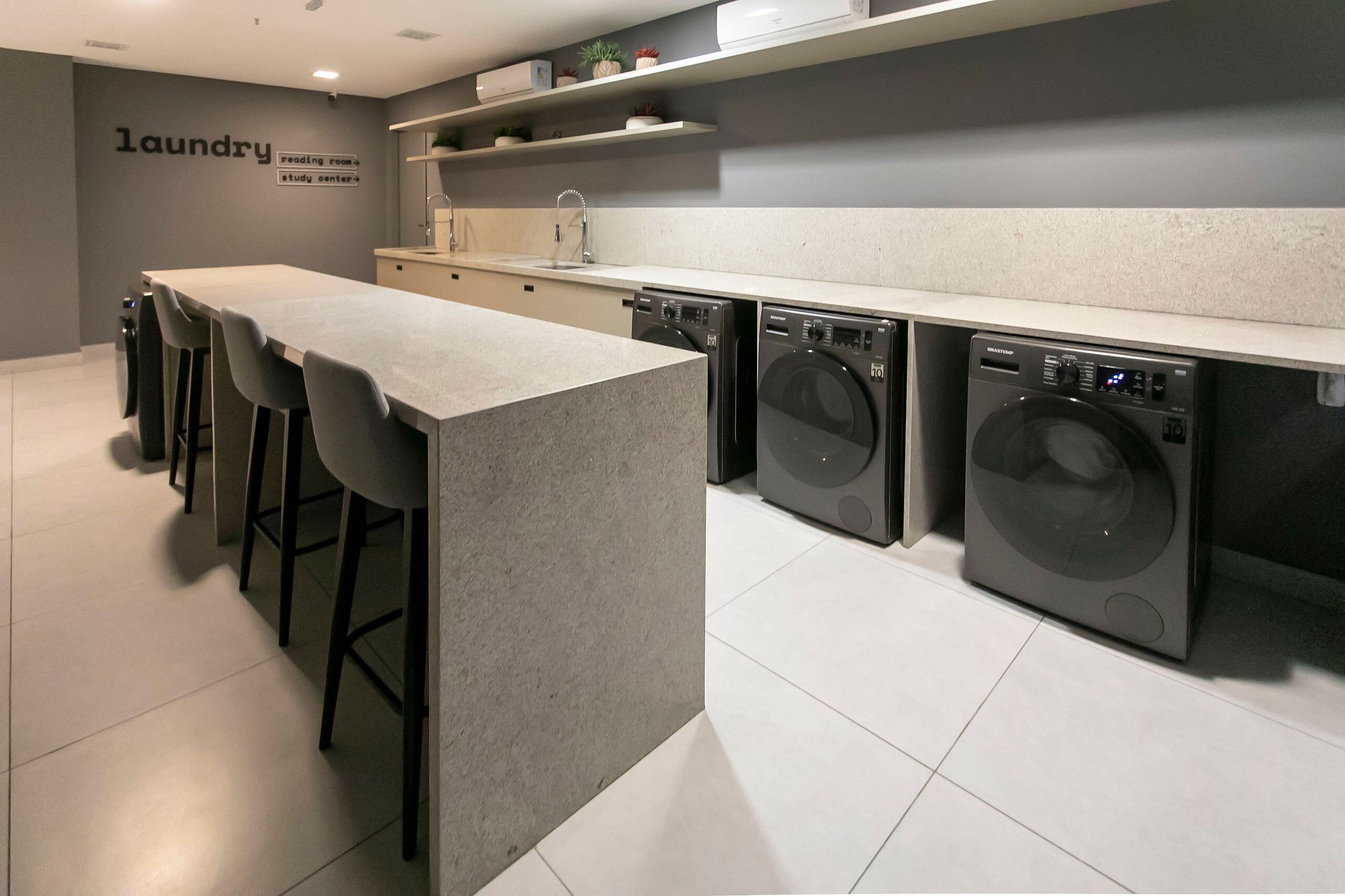 A modern laundry room with a long counter, bar stools, and two front-loading washing machines.