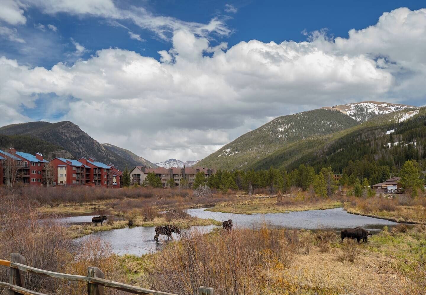 A mountainous landscape with a river, a wooden fence, and a row of red buildings with multiple floors.