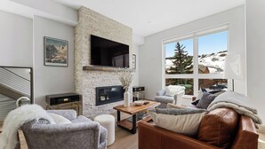A modern living room with a stone fireplace, a flat-screen TV, and a large window offering a view of a snowy landscape. - Contemporary luxury in the heart of Silverthorne (Silverthorne)