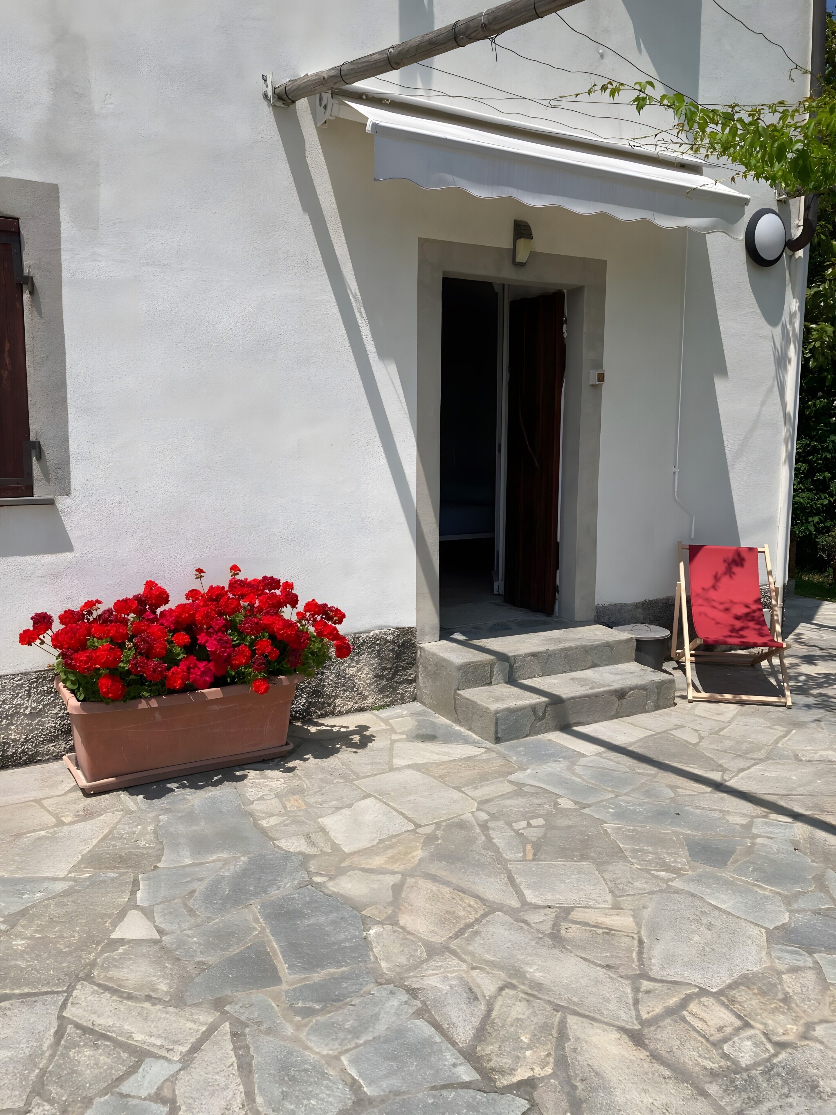 Un bâtiment à la façade blanche, avec une entrée pavée de pierres, un jardinier rouge et une chaise en bois.