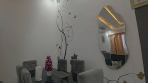 Un coin repas avec une table en verre, des chaises à revêtement gris, un mur décoratif orné de motifs abstraits et un miroir qui reflète une partie de la pièce.