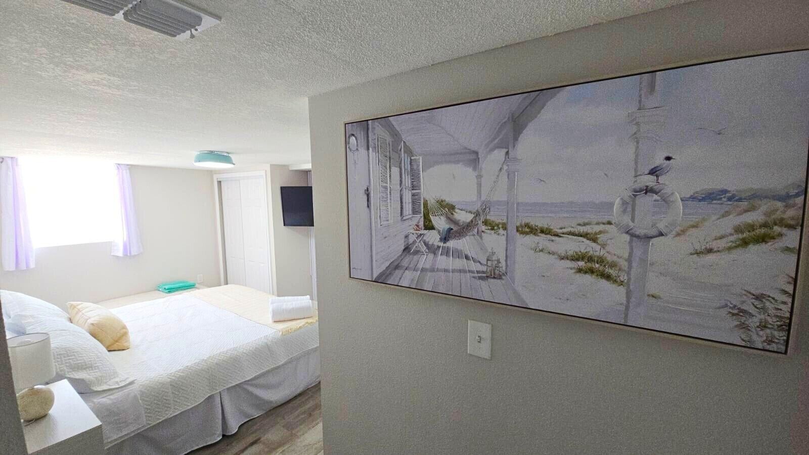 A bedroom with a bed, a wall-mounted TV, and a painting of a beach scene.