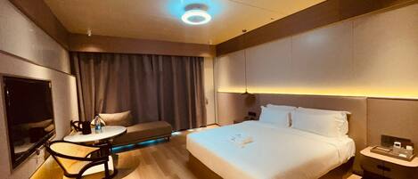A modern hotel room with a large bed, a desk with a chair, a TV, and a minibar.
