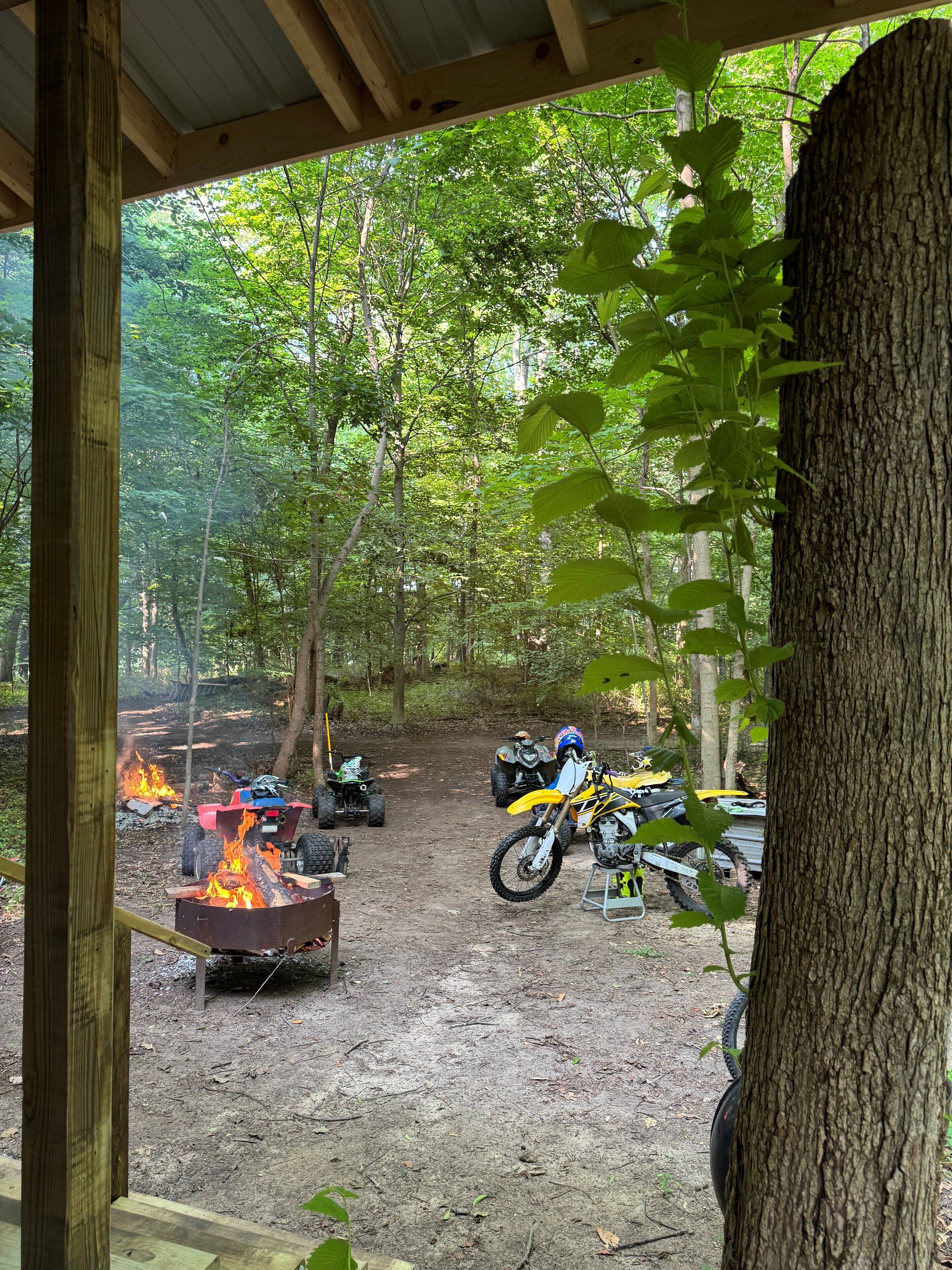 A forested area with dirt paths and ATVs parked near a fire pit.