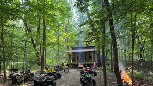 A forested area with a dirt path leading to a small cabin. There are several ATVs parked along the path, and a fire burning in the distance. - The Mill Creek Hideout (Mill Creek)