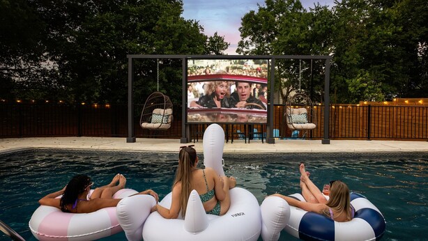 A pool area with inflatable loungers and a large screen displaying a movie.