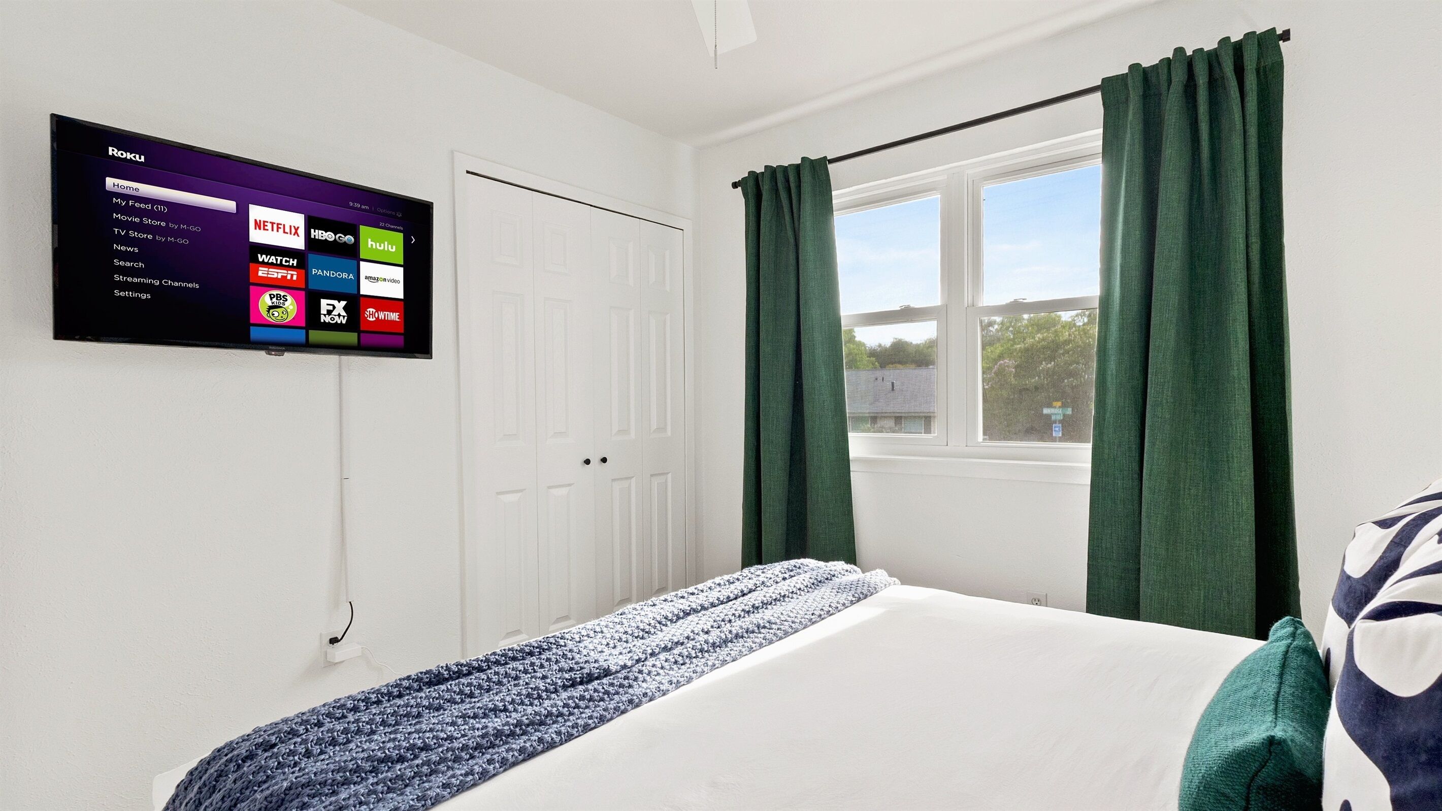 A hotel room with a bed, a TV mounted on the wall displaying streaming services, a window with curtains, and a view of the outside.
