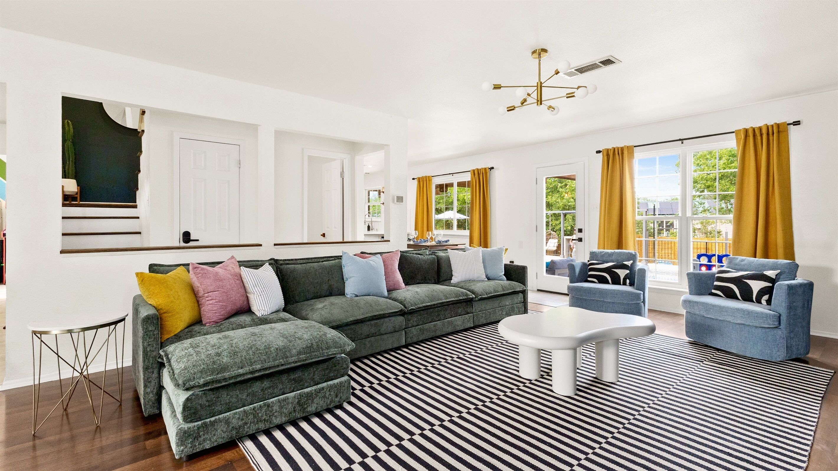 A modern living room with a sectional sofa, a coffee table, and two armchairs. There is a large window with curtains, a ceiling light fixture, and a black and white striped rug.