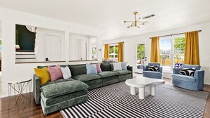 A modern living room with a sectional sofa, a coffee table, and two armchairs. There is a large window with curtains, a ceiling light fixture, and a black and white striped rug.