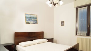 A bedroom with a wooden bed, a nightstand, a small table, a painting on the wall, and a window with curtains.