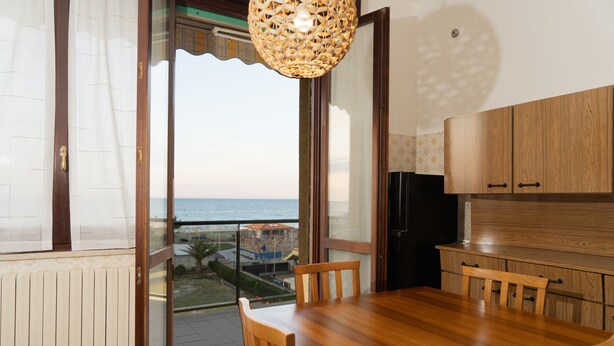 A balcony with a view of the sea, a wooden dining table, and a kitchen area.