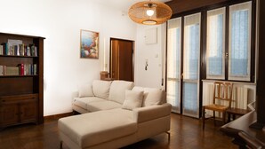 A living room with a sectional sofa, a wooden bookshelf, a wicker ceiling light, and a large window with blinds.