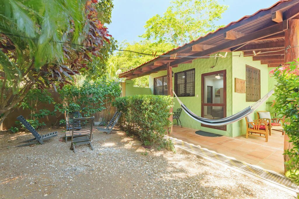 Una casa con fachada verde, una hamaca, una silla de madera y un pequeño patio.