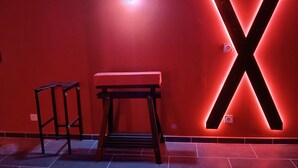 A room with a red wall, a large illuminated 'X' sign, a wooden bench, and a small table. - la chambre des plaisirs secrets Manduel (Manduel)