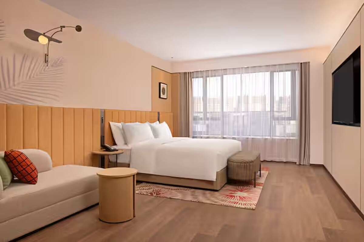 Habitación de hotel con cama, sofá, mesita de noche, lámpara y un ventanal con cortinas.