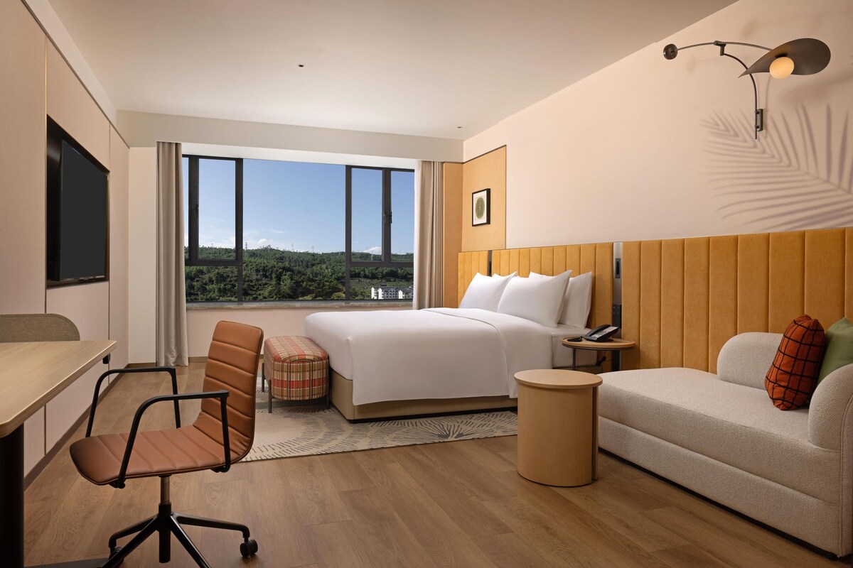 Habitación de hotel moderna con una cama grande, un sofá, un escritorio y una silla. Hay una ventana con vistas a zonas verdes.