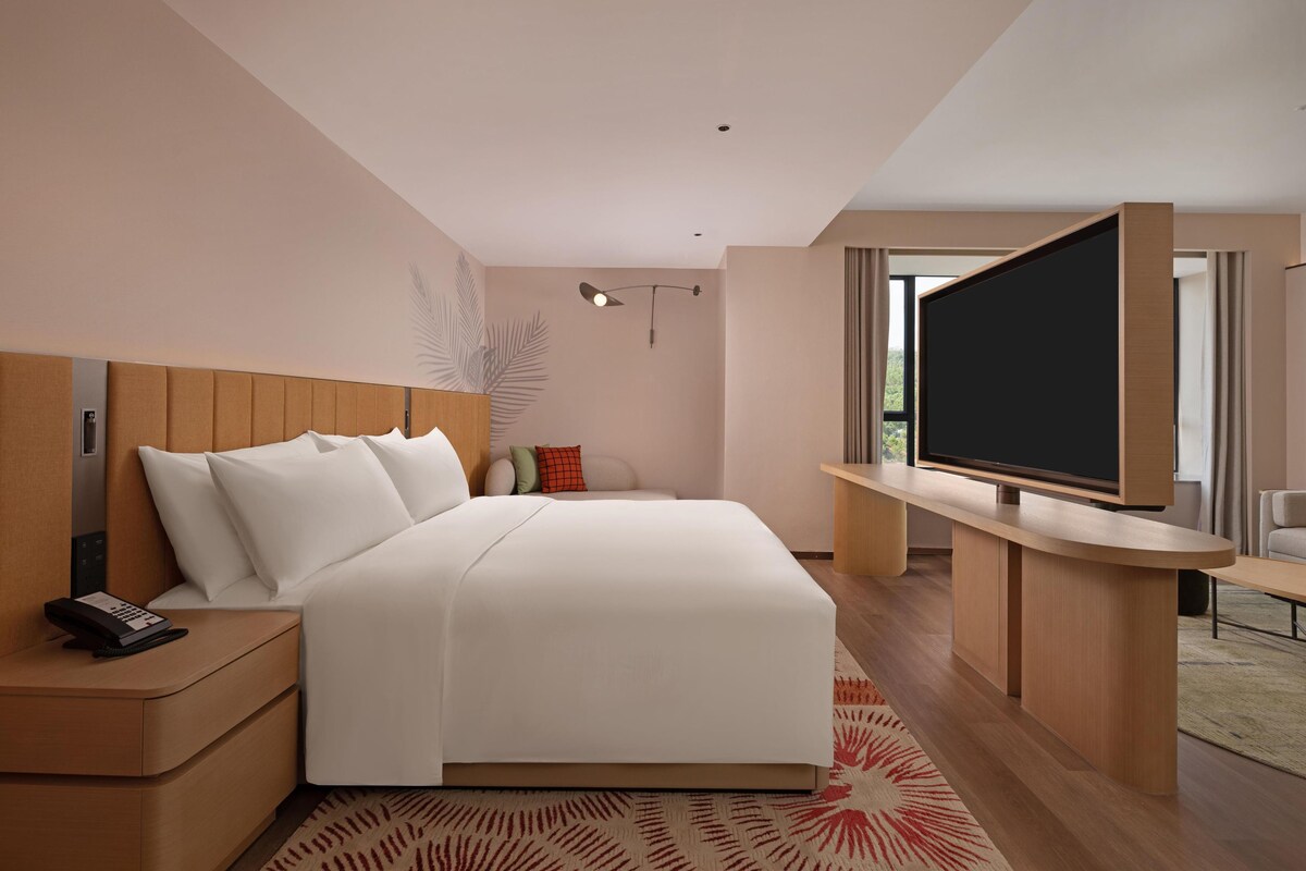 A modern hotel room with a large bed, a bedside table with a phone, a flat-screen TV mounted on a wooden console, and a large window offering a view of the outdoors.