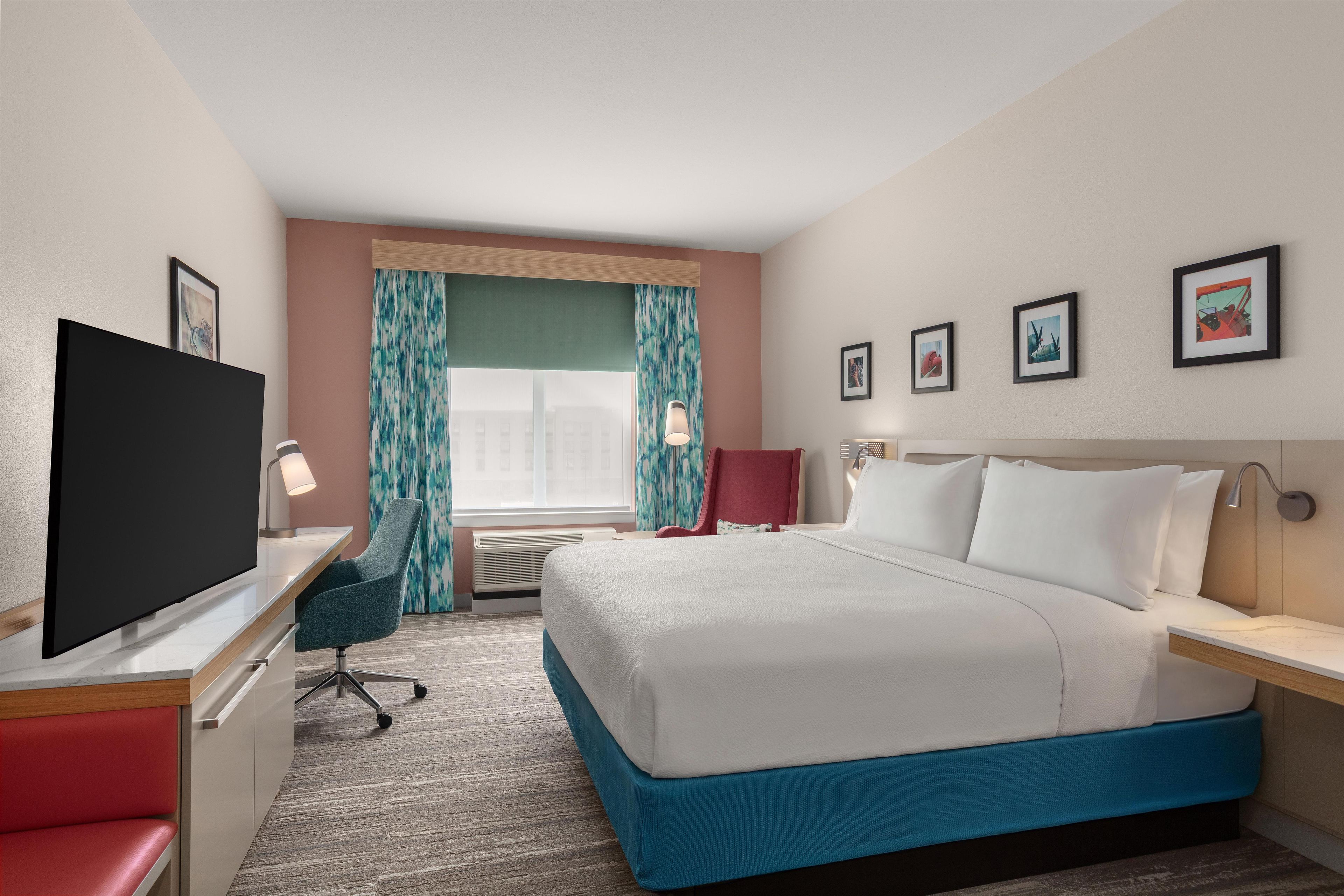 A hotel room with a bed, a desk with a TV, a chair, a red sofa, and a window with curtains.