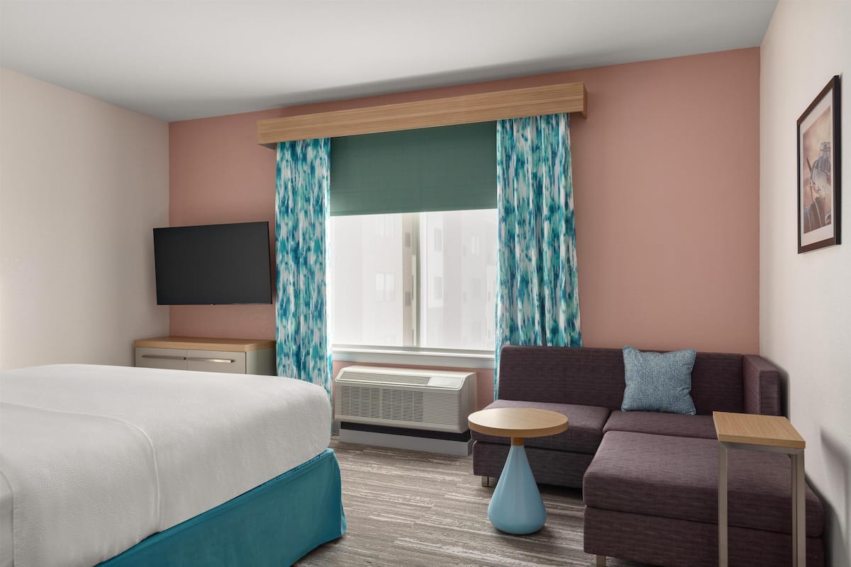 Una habitación de hotel con cama, sofá, una mesita, televisión y ventana con cortinas.