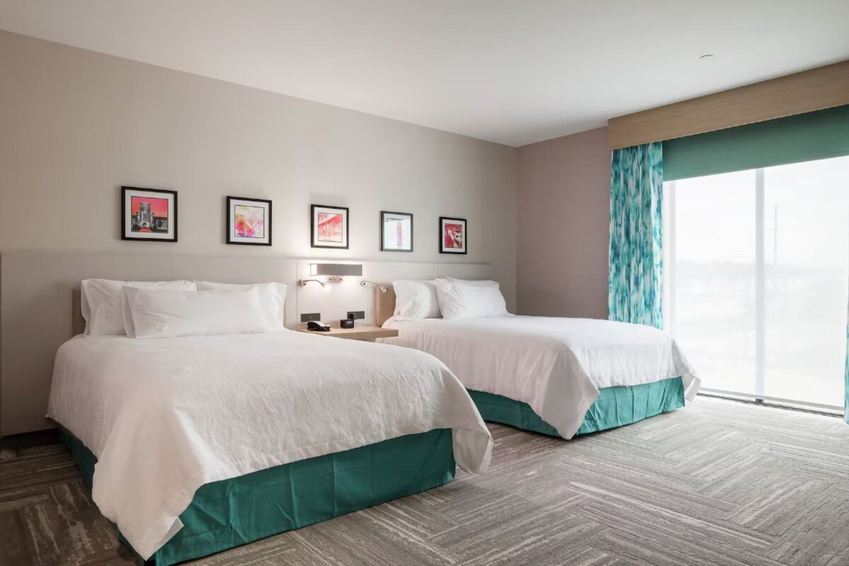 A hotel room with two beds, a nightstand, a lamp, and a window with curtains.